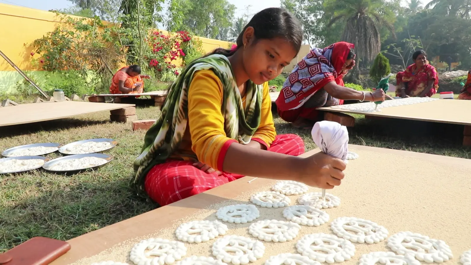 A young woman piping gohona bori onto a board of poppy seeds, with women at work behind her in a village garden