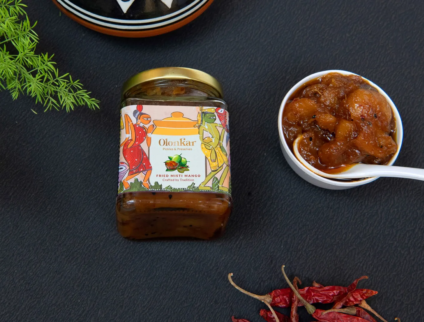 A jar of Olonkar fried misti mango beside a bowl of it, with dried red chillies
