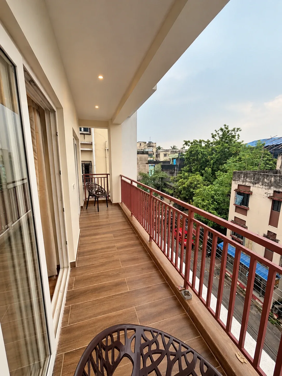 A long balcony with a wooden floor and red railings, over a leafy Kolkata lane