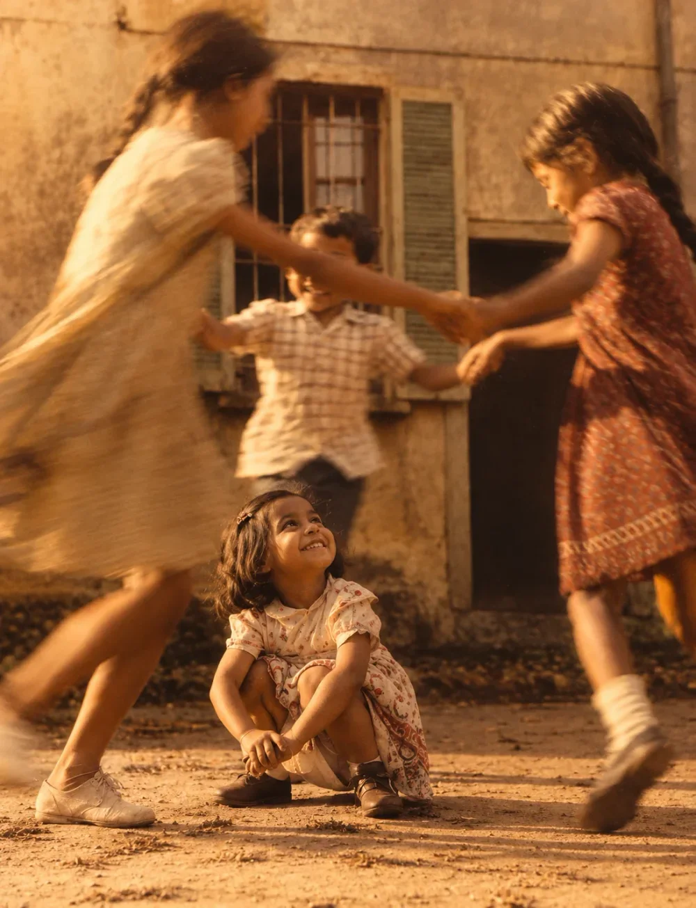 Girls playing ring-a-ring in a sunlit courtyard while the youngest sits and looks up, smiling